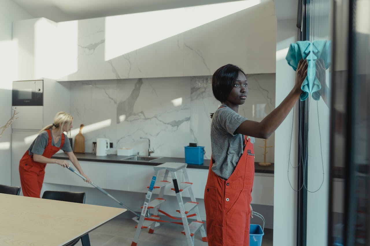 gallery-1 Two women cleaning a modern kitchen, wiping glass surfaces and mopping the floor.